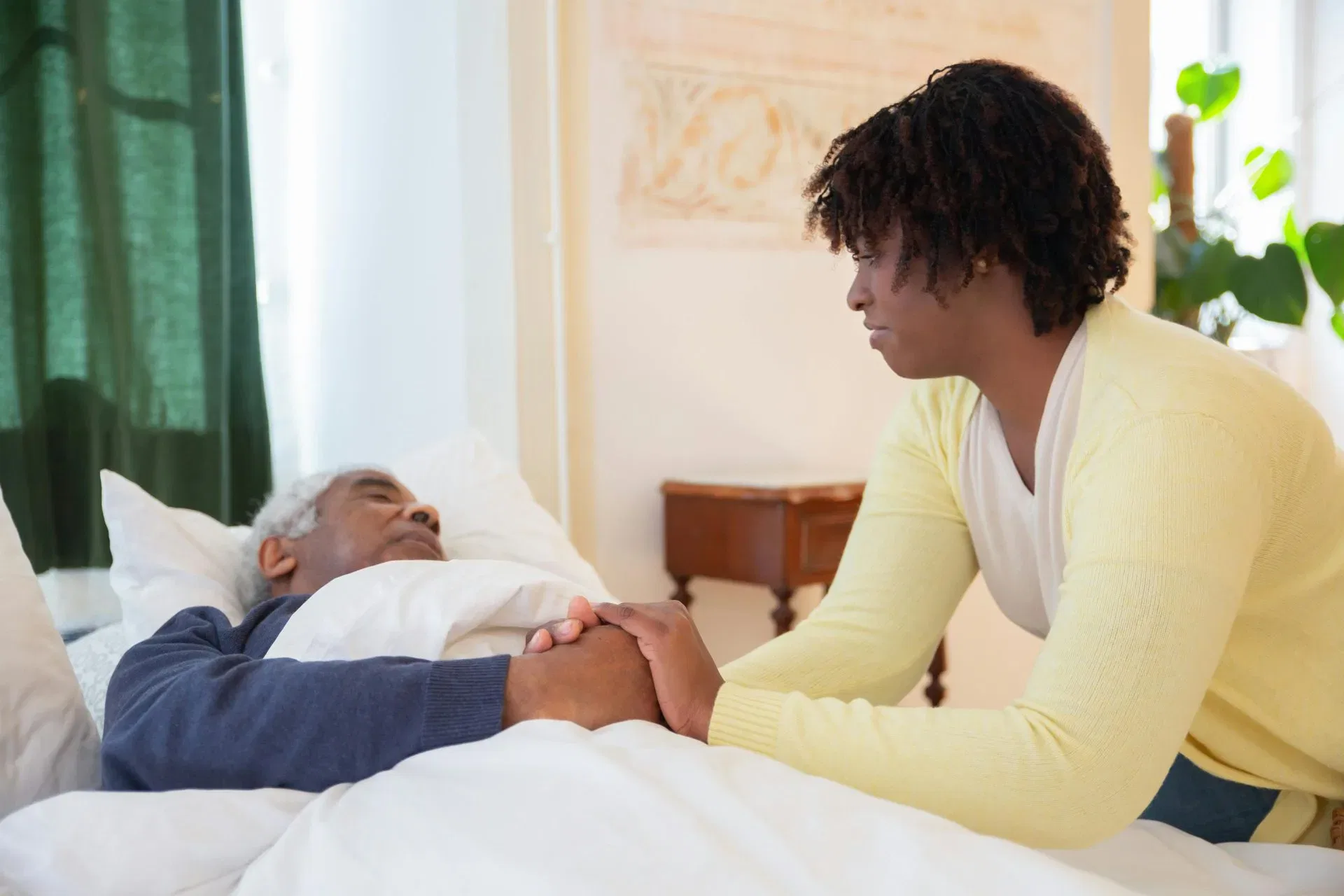 Woman in a yellow sweater holding their elderly relative's hand in bedroom, emphasizing guardianship and care